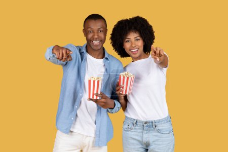Foto de Alegre sonriente pareja afroamericana en ropa casual sosteniendo cajas de palomitas de maíz a rayas rojas y blancas, señalando juguetonamente a la cámara con una expresión alegre, estudio - Imagen libre de derechos