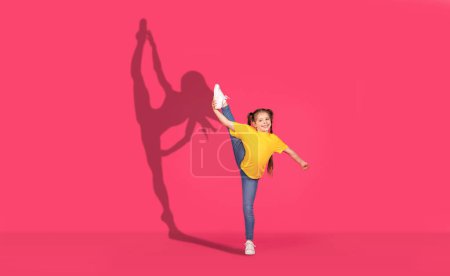 Téléchargez les photos : Une jeune fille joyeuse dans une chemise jaune et un jean effectue un coup de pied à la jambe haute avec enthousiasme, son ombre dynamique dansant sur un fond rose vif, représentant l'énergie et le bonheur - en image libre de droit