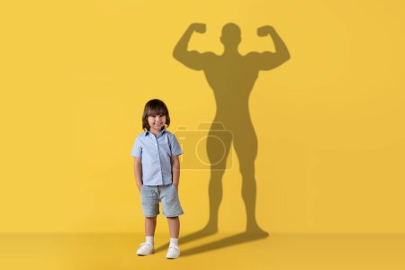 Téléchargez les photos : Un petit enfant caucasien joyeux se tient souriant avec une ombre de super-héros musclé et confiant jeté sur un fond jaune vif, symbolisant l'aspiration et le potentiel. Imagination, avenir - en image libre de droit