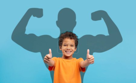 Photo for An exuberant young boy in an orange t-shirt gives two thumbs up, with a muscular superhero shadow behind him on a blue background, symbolizing strength and positivity. Imagination, future - Royalty Free Image