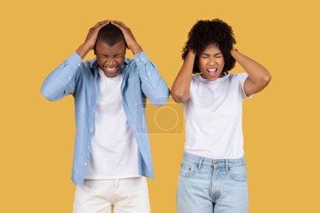 Photo for Stressed sad black young couple with hands on their heads, eyes closed, and faces contorted in frustration, standing against a unified mustard yellow background, studio. Stress - Royalty Free Image