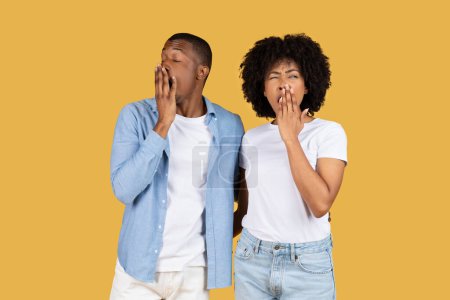 Photo for A young black couple in casual clothes looking tired and bored, yawning with hands over their mouths, standing against a plain mustard yellow background, studio. Sleep, late - Royalty Free Image