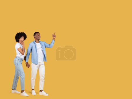 Téléchargez les photos : Heureux couple afro-américain millénaire ludique tenant la main, l'homme pointant vers le haut, souriant largement, alors que la femme regarde avec une expression curieuse, sur fond jaune vif - en image libre de droit