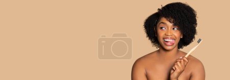 Téléchargez les photos : Joyeuse femme noire tenant une brosse à dents écologique, ludique et prête pour les soins dentaires, regardant l'espace libre, sur fond beige large, panorama - en image libre de droit