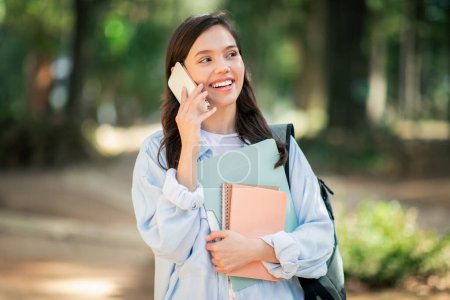 Cheerful european young woman student talking on a mobile phone while holding notebooks in a lush park, her expression reflecting a pleasant and engaging conversation, outdoor