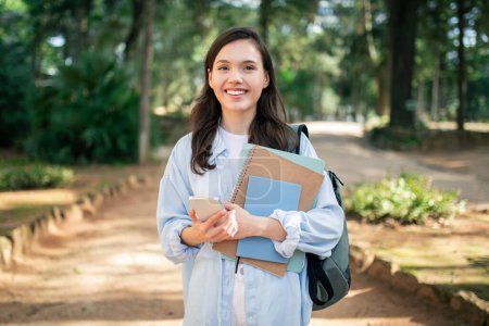 Radiant european young lady student holding books and a smartphone, looking optimistic amidst a natural park setting, embodying active academic life, outdoor. Study, education