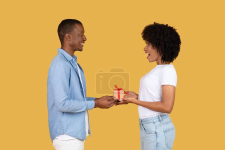 Photo for A young african american man in a light blue shirt happily giving a small, red-ribboned gift box to an excited woman in a white tee on a vibrant yellow background, studio - Royalty Free Image