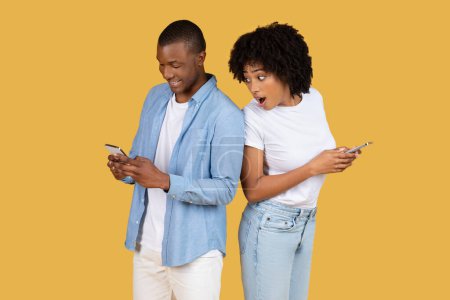 Photo for Surprised millennial African American woman peeking over the shoulder of a focused man, both using smartphones, hinting at a private or intriguing discovery on a yellow background - Royalty Free Image