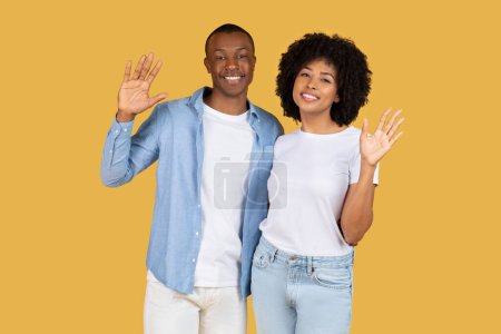 Foto de Feliz pareja afroamericana milenaria saludando a la cámara con sonrisas brillantes, casualmente vestida, creando un gesto amistoso y acogedor sobre un fondo amarillo - Imagen libre de derechos