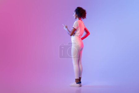 Téléchargez les photos : Femme noire focalisée en blanc pointant vers un écran invisible, imitant l'interaction à l'écran tactile, sur fond dégradé rose à bleu, espace libre - en image libre de droit