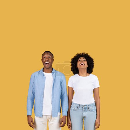 Photo for Amazed millennial African American man and woman in casual attire look upwards with open-mouthed wonderment, expressing surprise and anticipation against a solid yellow backdrop - Royalty Free Image