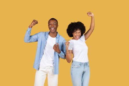 Téléchargez les photos : Couple afro-américain enthousiaste célébrant le succès avec les bras levés et les poings serrés, tous deux vêtus de vêtements décontractés avec des expressions joyeuses sur un fond jaune - en image libre de droit