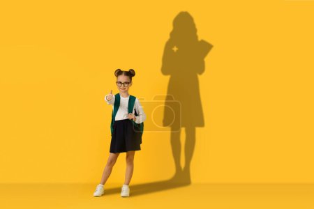 Photo for A confident schoolgirl with glasses and hair buns gives a thumbs up while holding a green backpack, her shadow on a yellow wall projecting a figure of success and academic achievement - Royalty Free Image