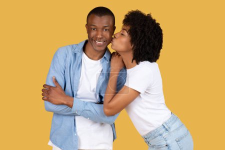 Foto de Afectuosa mujer afroamericana milenaria besa a un hombre sonriente en la mejilla, que cruza sus brazos con una expresión complacida, tanto en ropa casual sobre un fondo amarillo - Imagen libre de derechos