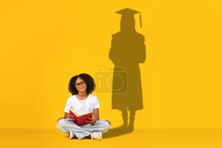 Téléchargez les photos : Une jeune fille studieuse s'assied jambes croisées en lisant un livre, avec son ombre sur un fond jaune illustrant un diplômé en casquette et robe, symbolisant la réussite scolaire future - en image libre de droit