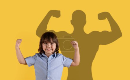 Téléchargez les photos : Un enfant souriant dans une chemise bleue montre les muscles avec une ombre de super-héros en arrière-plan, transmettant un message de force, de confiance et de puissance de l'imagination sur une toile de fond jaune - en image libre de droit