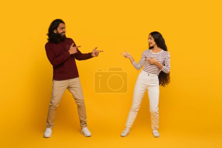Man and woman in casual attire playfully point at each other on a vivid yellow background