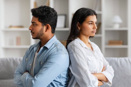 Upset young hindu couple sitting back-to-back on sofa, displaying signs of disagreement with arms crossed and serious expressions in light living room