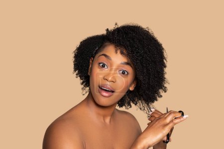 Photo for Young black woman with an expression of surprise with scissors in hand, cuts her voluminous curly hair, set against soft beige backdrop - Royalty Free Image