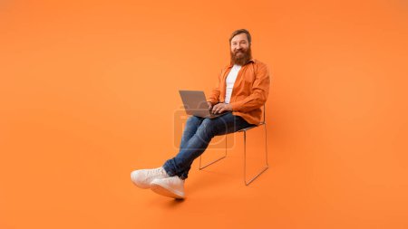 Téléchargez les photos : Casual redhaired homme d'âge moyen pigiste navigue sur son gadget d'ordinateur portable, mettant l'accent sur la technologie moderne et le travail indépendant en ligne, assis dans une chaise dans un cadre de studio orange. Panorama - en image libre de droit