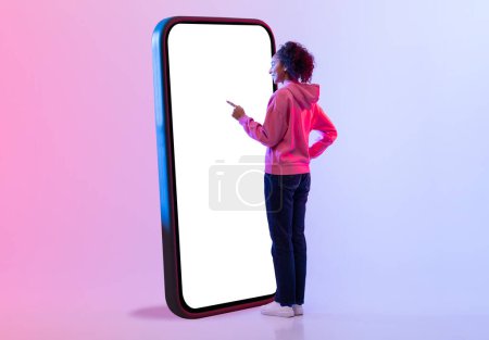 Téléchargez les photos : Vue latérale de la jeune femme aux cheveux bouclés, portant un sweat à capuche rose, s'engageant avec un énorme écran de smartphone en studio avec fond dégradé rose à bleu, maquette - en image libre de droit