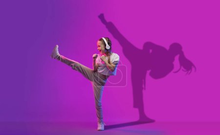 Foto de Chica joven feliz con auriculares patea alegremente alto en el aire, su silueta proyecta una sombra de baile sobre un fondo púrpura, mostrando su espíritu enérgico y pasión por la música y el movimiento - Imagen libre de derechos