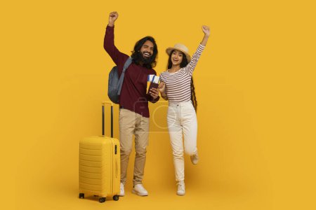 Joyful couple with luggage celebrating travel plans arms raised on a vivid yellow background