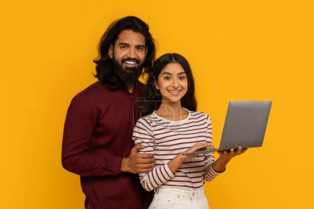 Couple with a laptop and a bright expression, considering potential travel plans on yellow backdrop