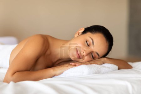 Téléchargez les photos : Jeune femme européenne détendue se reposant après un massage du corps cicatrisant, profitant de la détente à l'intérieur du spa de luxe, allongée les yeux fermés sur une table de massage dans un salon de beauté moderne, attendant la procédure - en image libre de droit