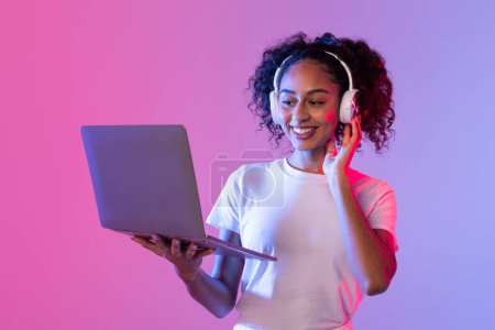 Photo for Woman in white T-shirt and jeans using a laptop and wearing headphones, set against a neon pink and purple background - Royalty Free Image