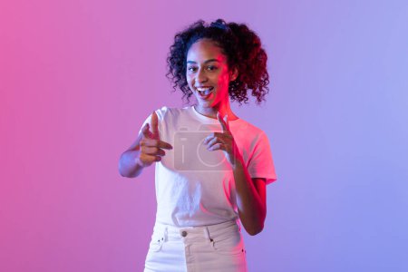 Photo for Excited woman pointing directly at the camera on a pink and purple neon-lit background, suggesting connection - Royalty Free Image
