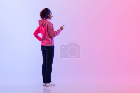 Photo for Rear view of young woman with curly hair and pink hoodie, pointing at virtual screen or pressing invisible button against soft pink and purple gradient background - Royalty Free Image
