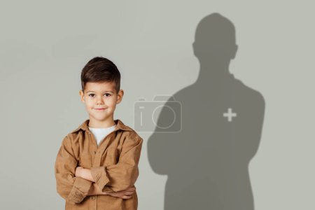 Téléchargez les photos : Petit garçon confiant, les bras croisés, souriant devant une toile de fond grise, son ombre projetant la silhouette d'un professionnel de la santé avec un signe plus, représentant potentiel et soin - en image libre de droit