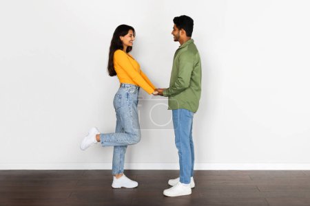 Foto de Sonriendo joven pareja hindú juguetonamente cogidas de la mano, mujer levantando su pierna en pose coqueta, de pie contra la pared blanca, mostrando momento romántico - Imagen libre de derechos