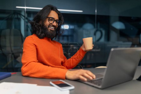 Content professional in orange knitwear sipping coffee during work hours in a vibrant office