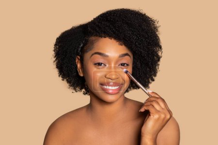 Photo for Glowing black woman with natural curls carefully applies concealer under her eye with makeup brush, enjoy beauty routine against beige backdrop - Royalty Free Image