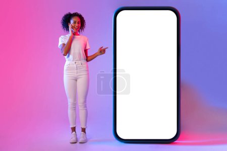 Photo for Surprised young black woman in white, interacting with huge blank smartphone display, presenting application or advertisement on neon dual-tone background - Royalty Free Image