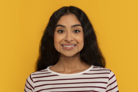 Téléchargez les photos : Une jeune femme en chemise rayée sourit en toute confiance dans un fond jaune vif, rayonnant de positivité - en image libre de droit
