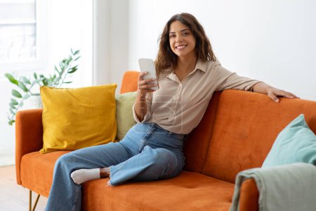 Foto de Mujer joven sonriente con teléfono inteligente en la mano sentada en el sofá y mirando a la cámara, hermosa hembra milenaria usando el teléfono móvil para la comunicación en línea o navegar por la nueva aplicación, relajarse en casa - Imagen libre de derechos