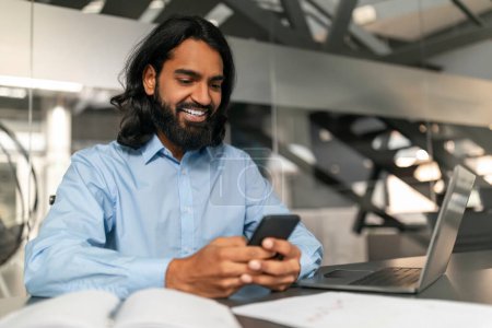 Glücklicher Geschäftsmann checkt Nachrichten auf seinem Smartphone im Büro, Laptop und Notebook auf seinem Schreibtisch