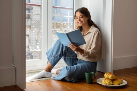 Photo for Smiling young woman reading interesting book while comfortably sitting on floor near window, beautiful female relaxing at home with cup of coffee and fresh croissants nearby, enjoying weekend - Royalty Free Image