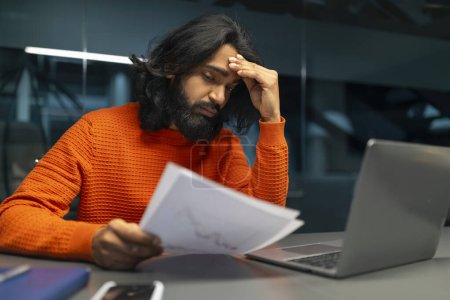 Stressed male looks at documents while showing signs of headache and stress at work