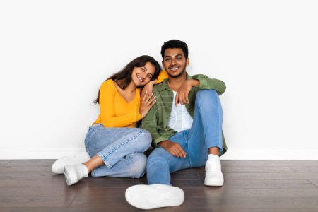 Téléchargez les photos : Couple indien affectueux en tenue décontractée assis étroitement sur le sol, femme appuyée sur l'épaule mans, souriant et se sentant content - en image libre de droit