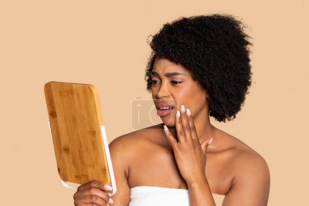 Téléchargez les photos : Femme noire préoccupée en serviette, inspectant son teint avec un miroir en bois, exprimant des soucis de santé de la peau, sur fond beige harmonieux - en image libre de droit