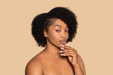Pensive black woman with natural curls gently rests hand on chin, expressing contemplation and elegance on neutral beige background
