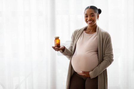 Cheerful pregnant young black woman embracing her big tummy, holding jar with pills drugs medication, standing next to window at home, copy space. Supplements for future moms
