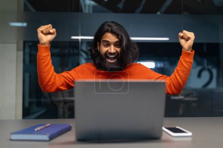 Photo for An elated man with arms up in triumph by the laptop in an office environment - Royalty Free Image