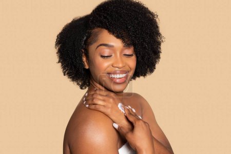 Photo for Cheerful young black woman with curly hair applies moisturizer on her hand, enjoying her skincare routine in front of beige background - Royalty Free Image