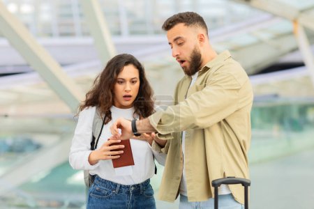 Photo for Stressed Young Couple Late For Flight, Upset Young Spouses Standing At Airport Terminal And Checking Time On Wristwatch, Holding Passports With Tickets, Suffering Problems While Travelling Together - Royalty Free Image