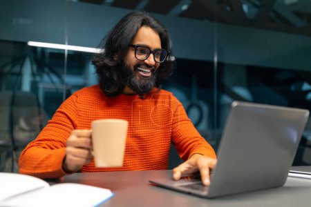 Photo for A man wearing glasses and an orange sweater holds a coffee mug while using a laptop, with a book on the table - Royalty Free Image